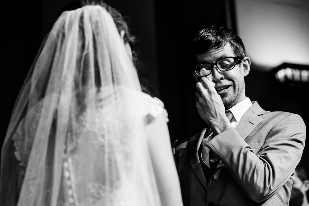 black and white image of the groom wiping away his tears as the bride looks at him during their wedding ceremony inside Grace Road Church