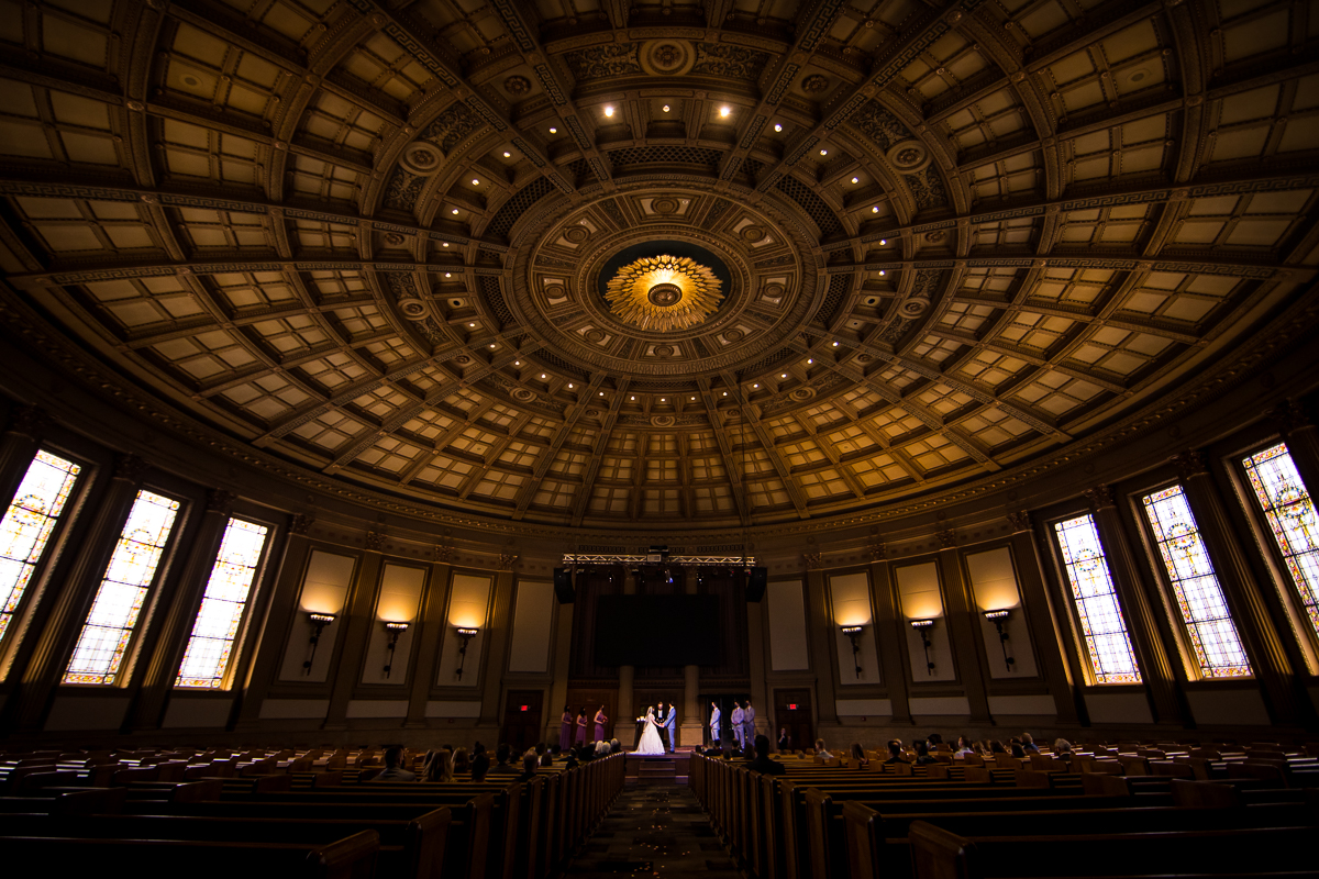 creative unique image of the bride and groom holding hands at the front of the church with the massive round ceiling the focus of the image for this Rochester NY wedding ceremony