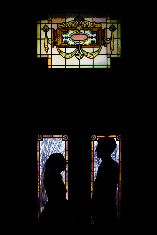 unique, creative photo of the bride and groom looking at one another as they stand in the stained glass windows and we see their silhouettes for their Rochester NY wedding