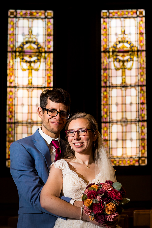 colorful, unique portrait of the bride and groom smiling at the camera with stained glass windows behind them after their Rochester NY wedding ceremony