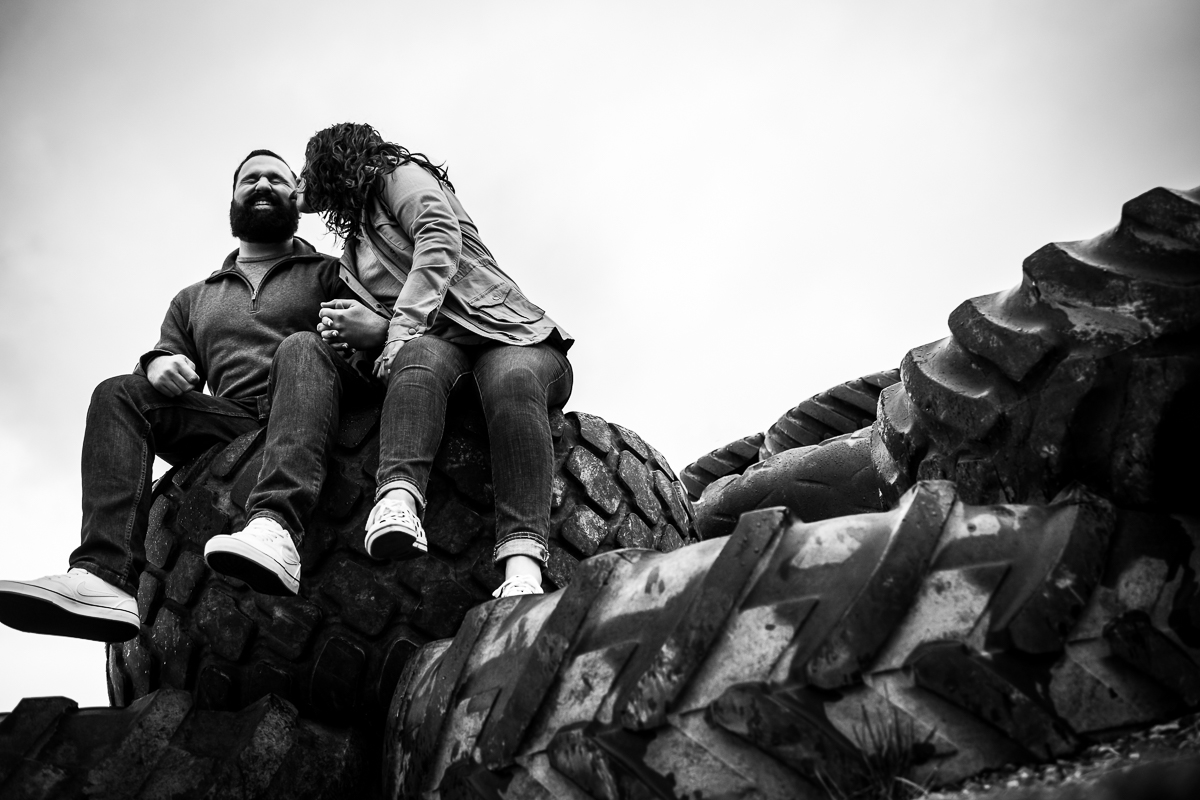 black and white image of the couple sitting on a pile of tires as they kiss one another during their rainy session in maryland
