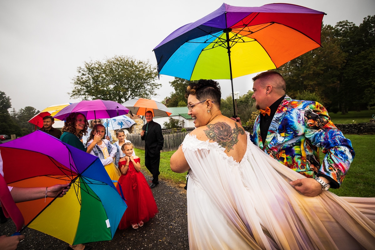 LGBTQ+ Wedding Photographer, Lisa Rhinehart captured a colorful, vibrant image of the of couple holding their rainbow umbrellas while they smile at their guests which are also holding rainbow umbrellas during their waynesboro pa backyard wedding