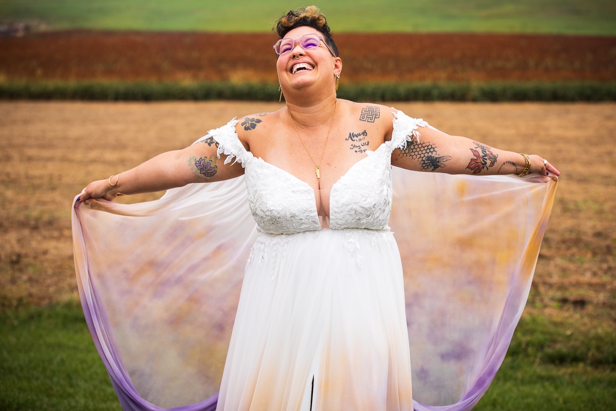 LGBTQIA+ Wedding Photographer, Lisa Rhinehart captured a vibrant colorful image of the bride with their arms holding out their unique purple, orange, and white dress for their waynesboro backyard preparation photos