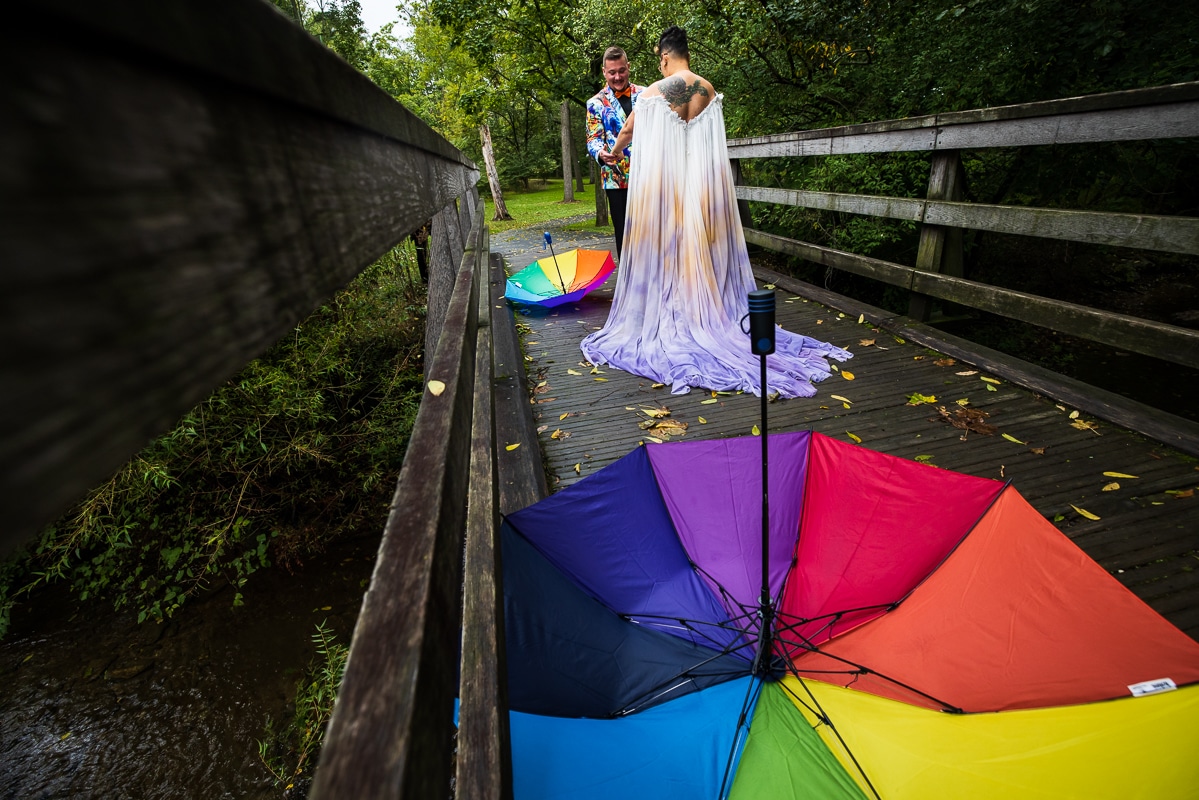 LGBTQ+ Wedding Photographer, Lisa Rhinehart captures the couple holding hands during their first look in their colorful, vibrant wedding attire while their rainbow umbrellas sit on the bridge behind them 
