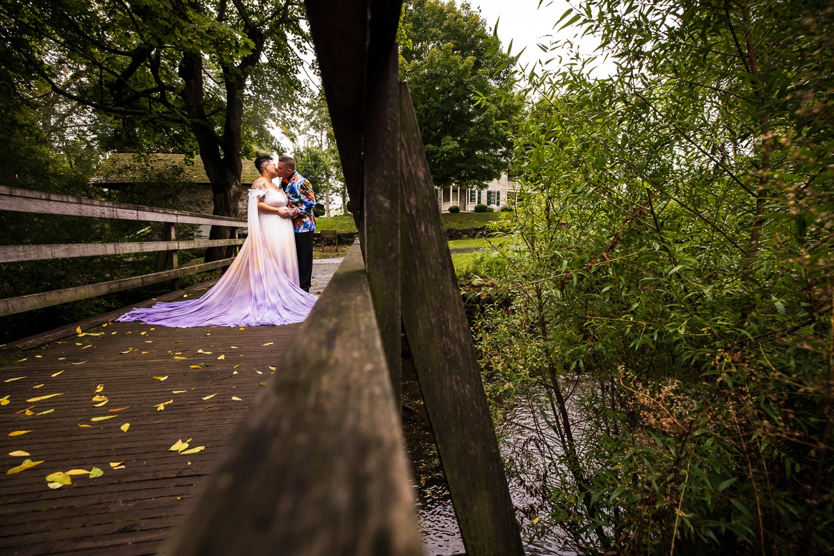 Unique creative colorful image of this queer and trans couple kissing in their colorful wedding attire on a bridge at Renfrew Park before their backyard wedding ceremony in Pennsylvania