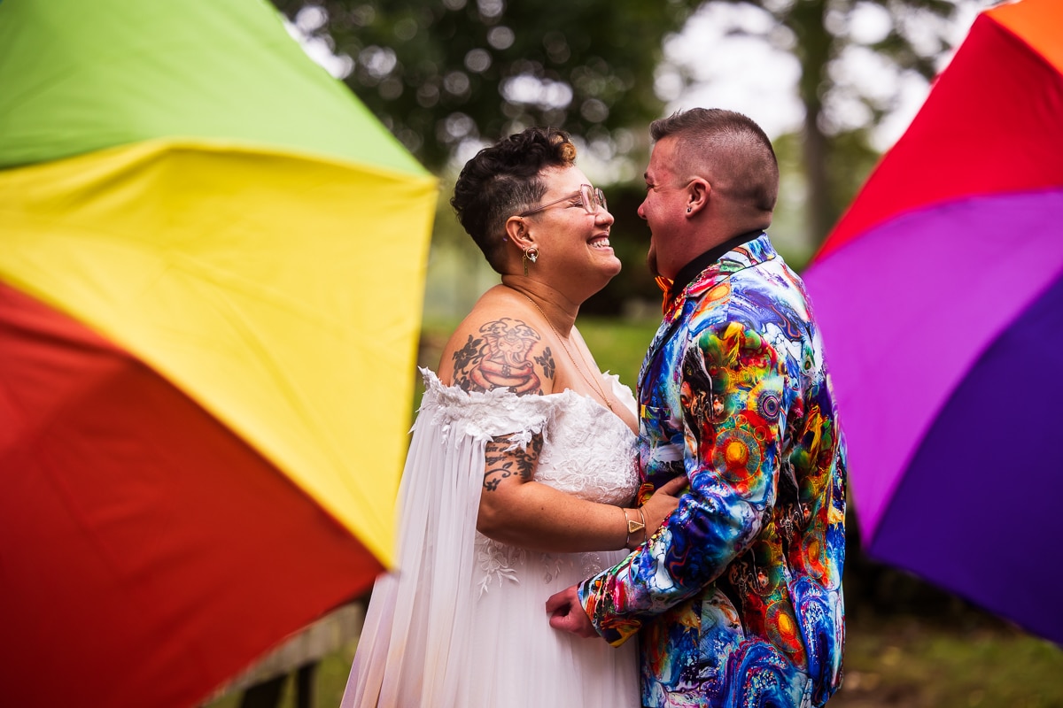 LGBTQ+ Wedding Photographer, Lisa Rhinehart captured this queer and trans couple hugging one another as they smile at each other with rainbow umbrellas in the background behind them before their backyard wedding in waynesboro, pa