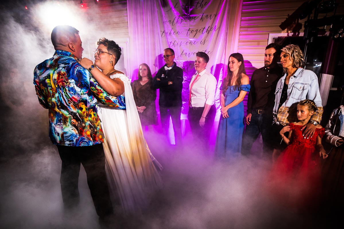 Colorful, vibrant, unique image of this queer and trans couple sharing their first dance as their friends and family watch from behind as fog comes up around them during their backyard wedding reception