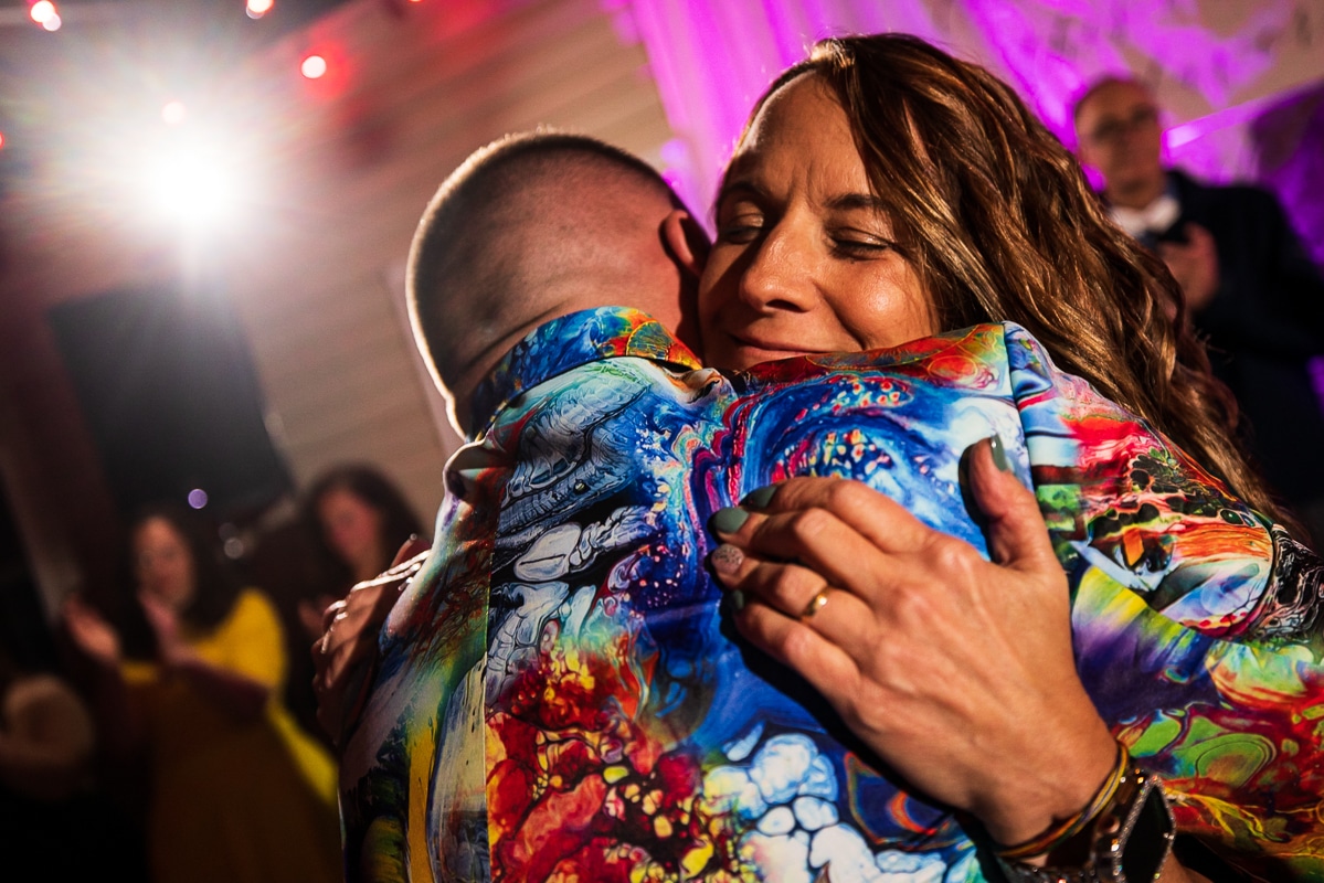 LGBTQIA+ Wedding Photographer, Lisa Rhinehart captures this special moment between the groom and his mom hugging during their parent dance as the mom has a huge loving smile on her face and she embraces the groom in his colorful unique wedding attire during this backyard wedding reception in waynesboro pa