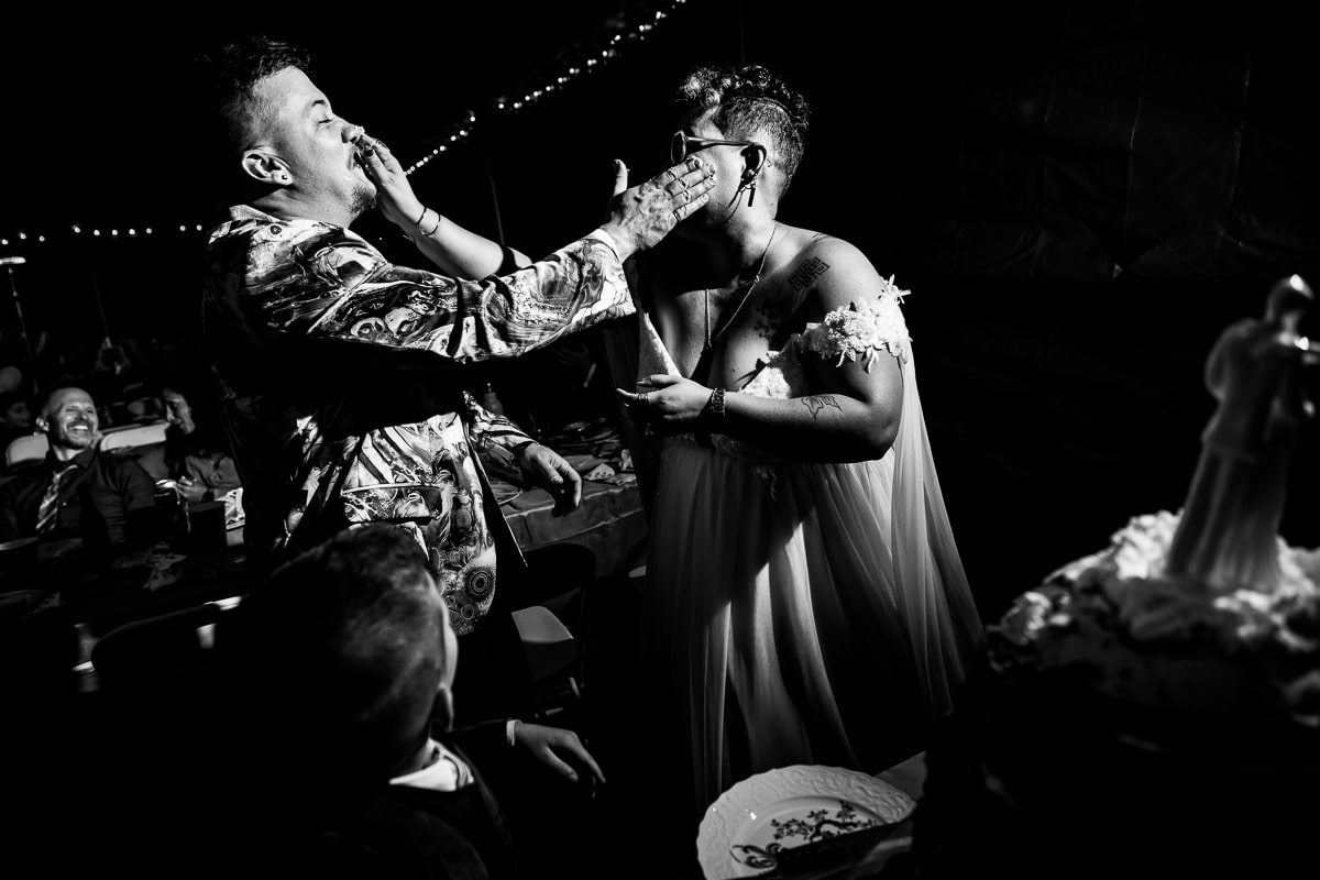 Black and white image of this queer and trans couple smashing their cookie cake into one another's faces during their backyard wedding reception 