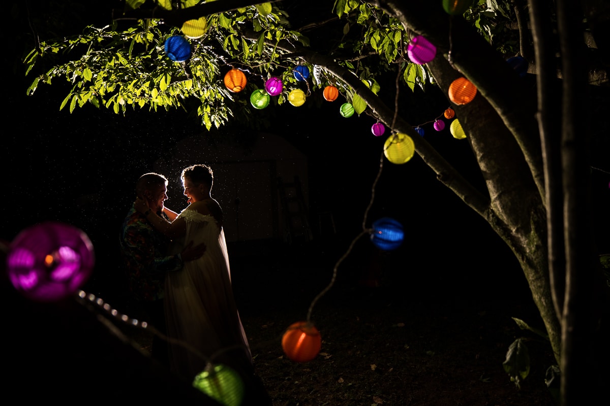 LGBTQIA+ Wedding Photographer, Lisa Rhinehart captures a unique, creative, colorful end of the night shot of the couple hugging one another as they are surrounded by trees and colorful lantern lights for their backyard wedding in waynesboro pa