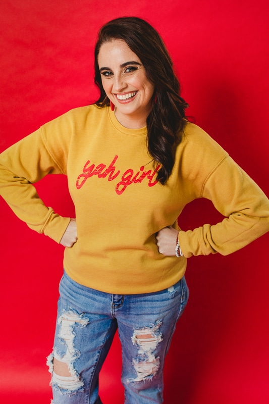 Lisa Rhinehart, owner of Rhinehart Photography, who is an award winning, creative photographer standing in front of a bright red background in a yellow sweatshirt as one of the Best wedding photographers in the USA