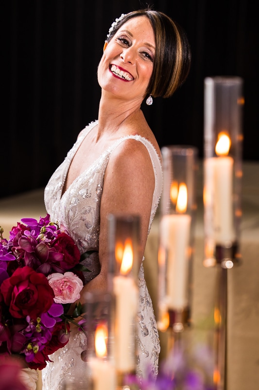 The bride is smiling towards the camera while holding her colorful bouquet with candles burning behind her before her wedding ceremony