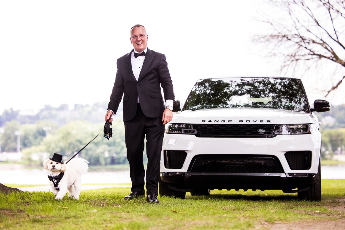 The groom and the couple's dog both dress in tuxedos walking towards the wedding venue with the Susquehanna River and a white range rover in the background for this Harrisburg Civic club wedding