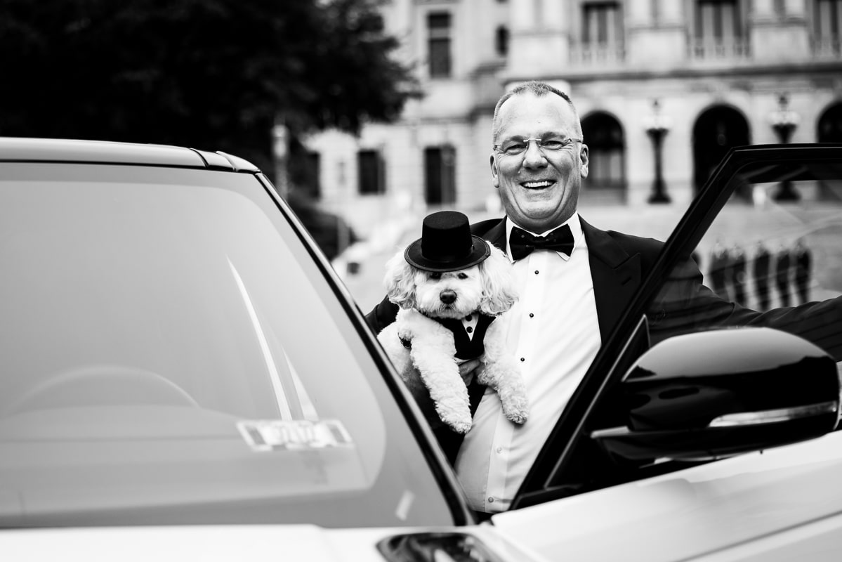 Black and white image of the groom holding the dog smiling as he gets out of the white range rover before their Harrisburg civic club wedding ceremony
