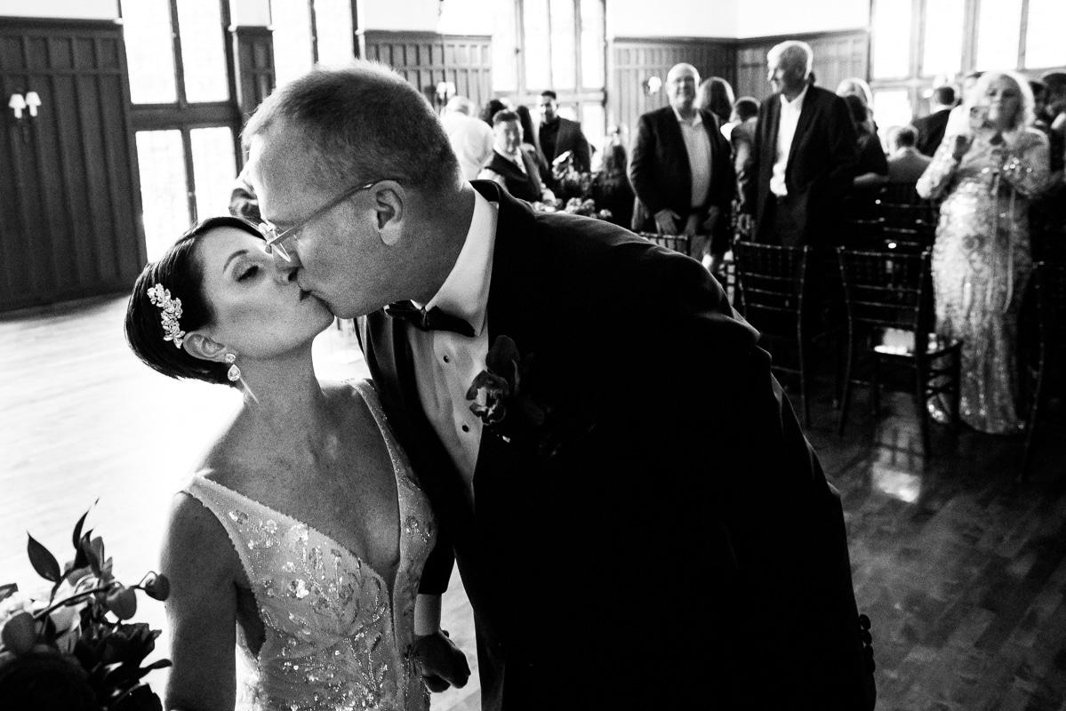 black and white image of the bride and groom kissing while their guests stand in the background watching