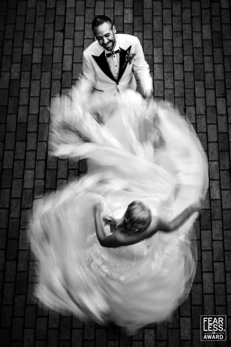 Black and white photo of the groom holding the brides dress as she spins around won another Fearless Photographers Award