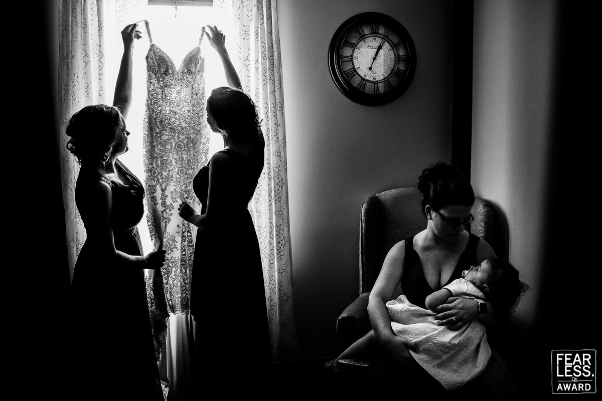 Black and white silhouetted image of two bridesmaids holding the wedding dress in the window with someone holding a baby in the lower half of the photograph