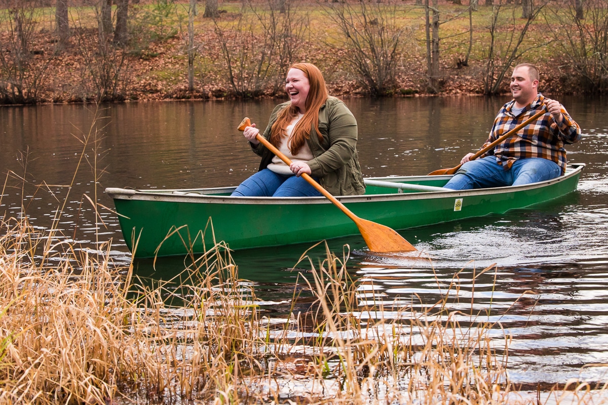 Image of the couple sitting, laughing and smiling in a green canoe paddling back to the side of the pond during their engagement session in central pa