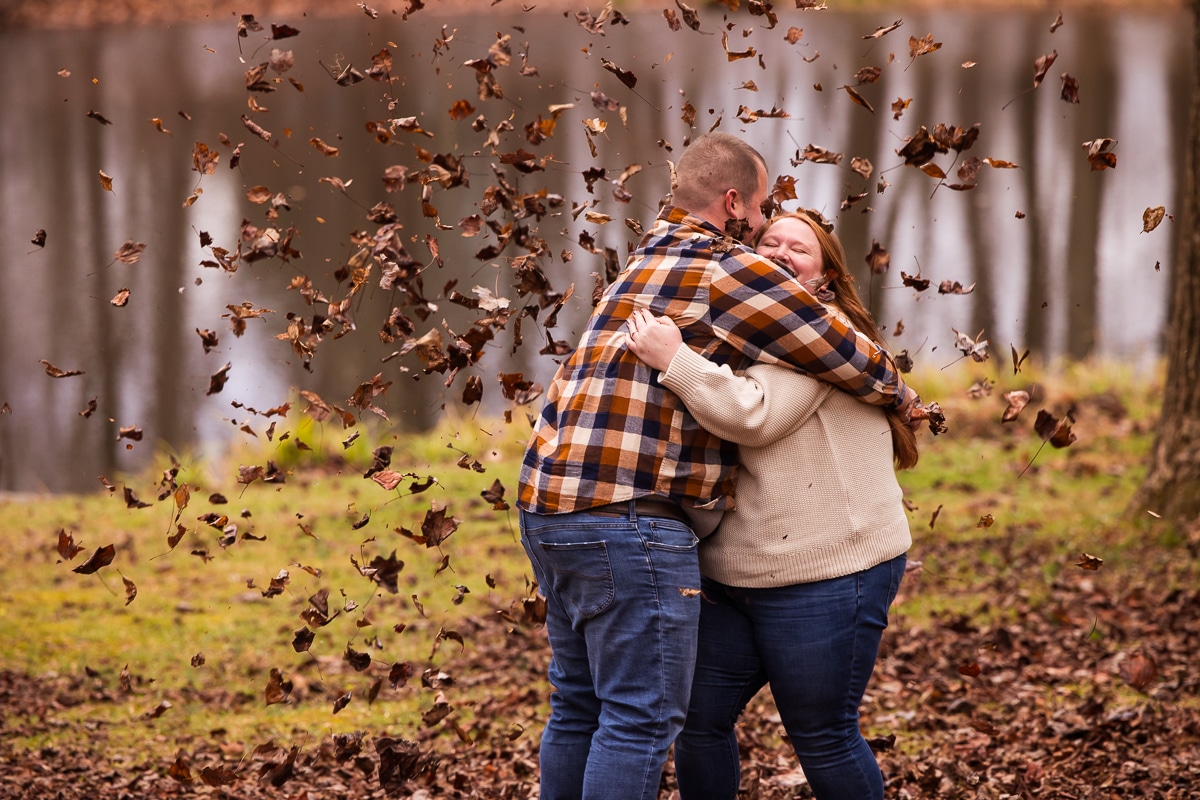Creative Engagement Photographer, lisa rhinehart, captures the couple as they hug one another with leaves flying around them and the large pond behind them 