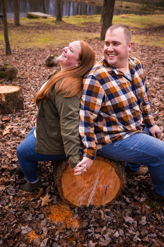 Image of the couple sitting on a cut down tree log laughing and smiling at each other surrounded by fallen leaves during their fall engagement session in central pa 