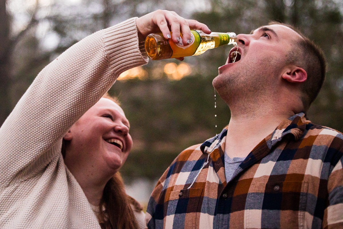 image of the future bride pouring beer into her fiances mouth as it drips down onto his shirt during their engagement session in pennsylvania