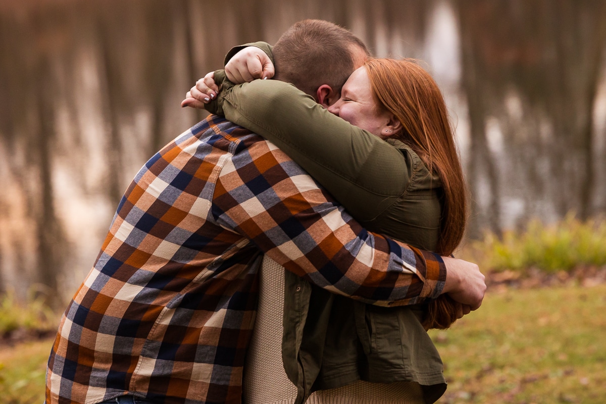 Creative Engagement Photographer, lisa rhinehart, captures the couple hugging and smiling one another during their outdoor fall engagement session with the pond behind them reflecting all of the trees 