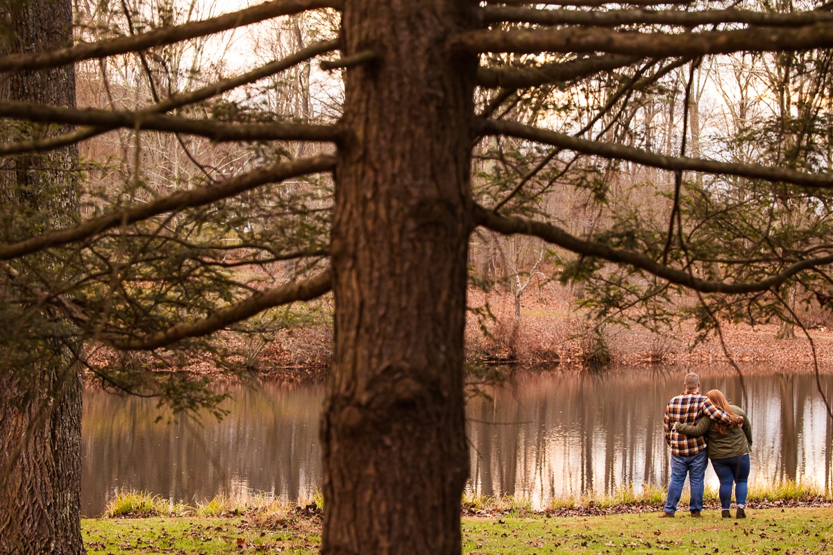 image of the couple standing together looking at the pond with their arms wrapped around one another with a giant tree in the foreground during their fall engagement shoot in central pa 