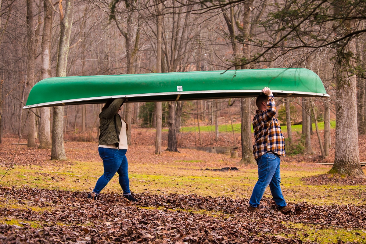 Image of the couple carrying a green canoe during their outdoor engagement session walking through all the fallen fall leaves on the grass