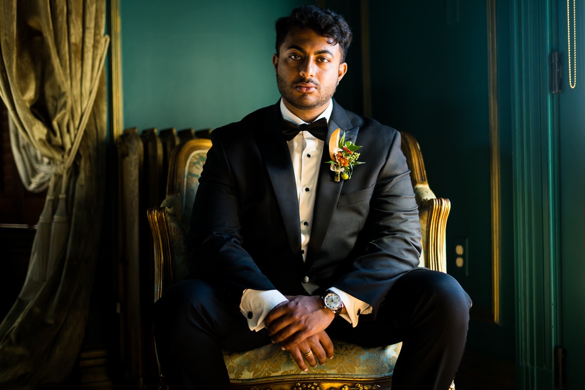 Image of the groom with a serious look on his face as he sits in front of a turquoise colored wall with color accents around him inside of the Willows at Ashcombe Mansion in Mechanicsburg PA