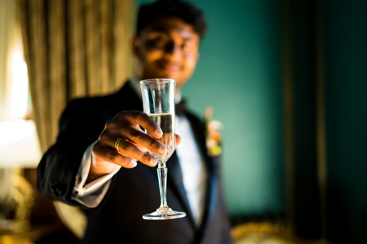 Unique, creative image of the groom out of focus as he holds a glass up towards the camera which is in focus as well as his ring against a vibrant turquoise backdrop with gold accents inside the mansion