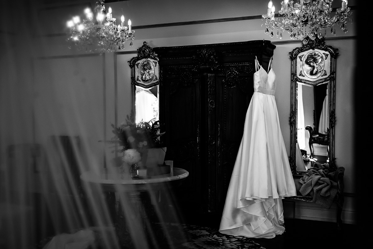 Black and white image of the bride's dress hanging from the furniture with chandeliers and mirrors decorating the inside of the room at the mansion 