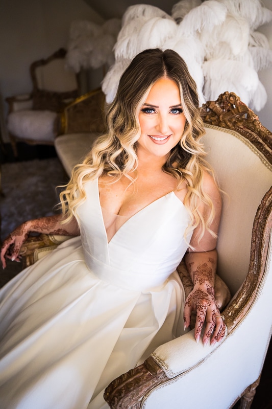 Light and airy image of the bride sitting in a white chair with wooden accents in her dress smiling up at the camera before her Ashcombe Mansion Wedding ceremony 