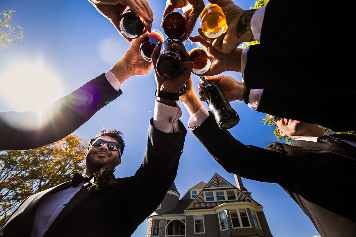 unique, creative perspective of the groom and his groomsmen holding drinks up in a circle with the ashcombe mansion in the background and a bright blue fall sky 