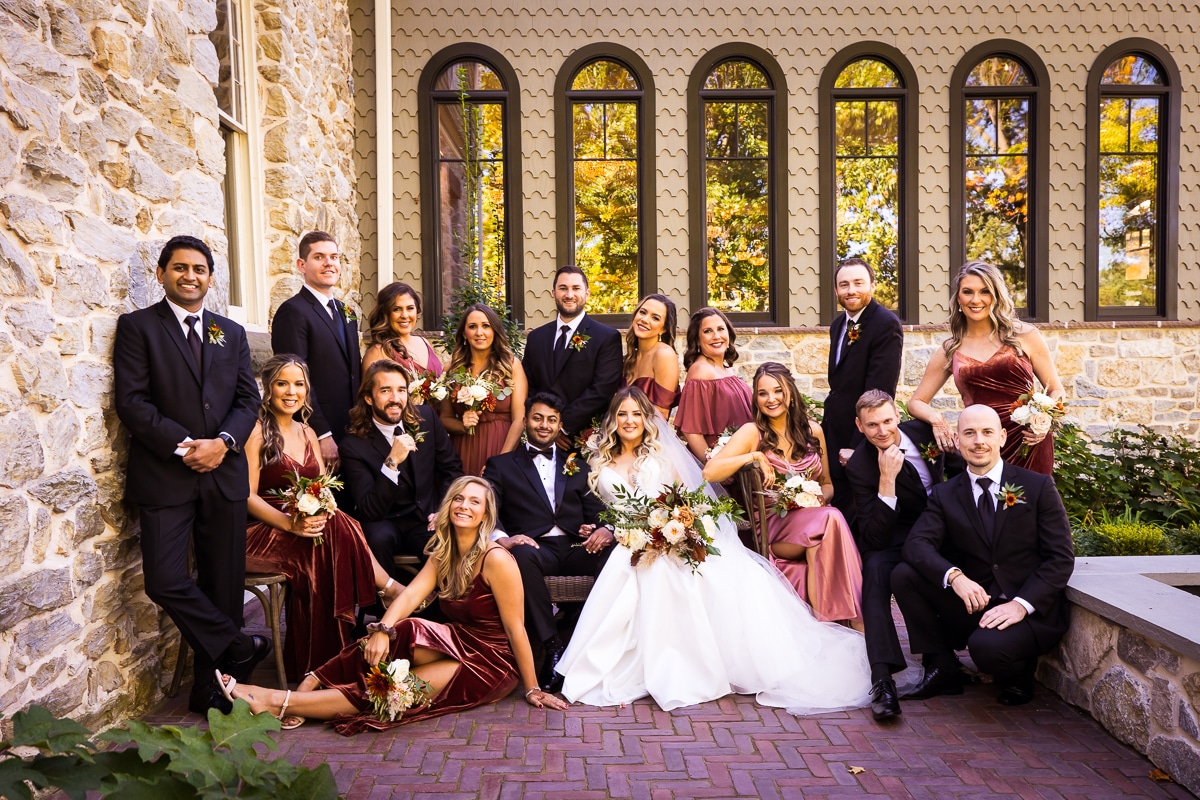 traditional wedding party portrait with everyone posed and smiling towards the camera in their black tuxedos and maroon dresses with their stone wall of the mansion and windows behind them in mechanicsburg pa 