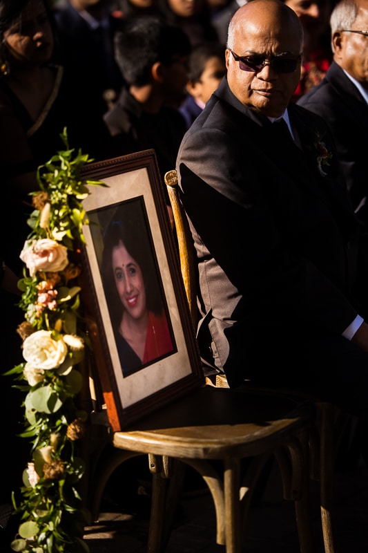 image of the father of the groom is looking down at a photo of his wife sitting on a chair at their Ashcombe Mansion wedding ceremony 