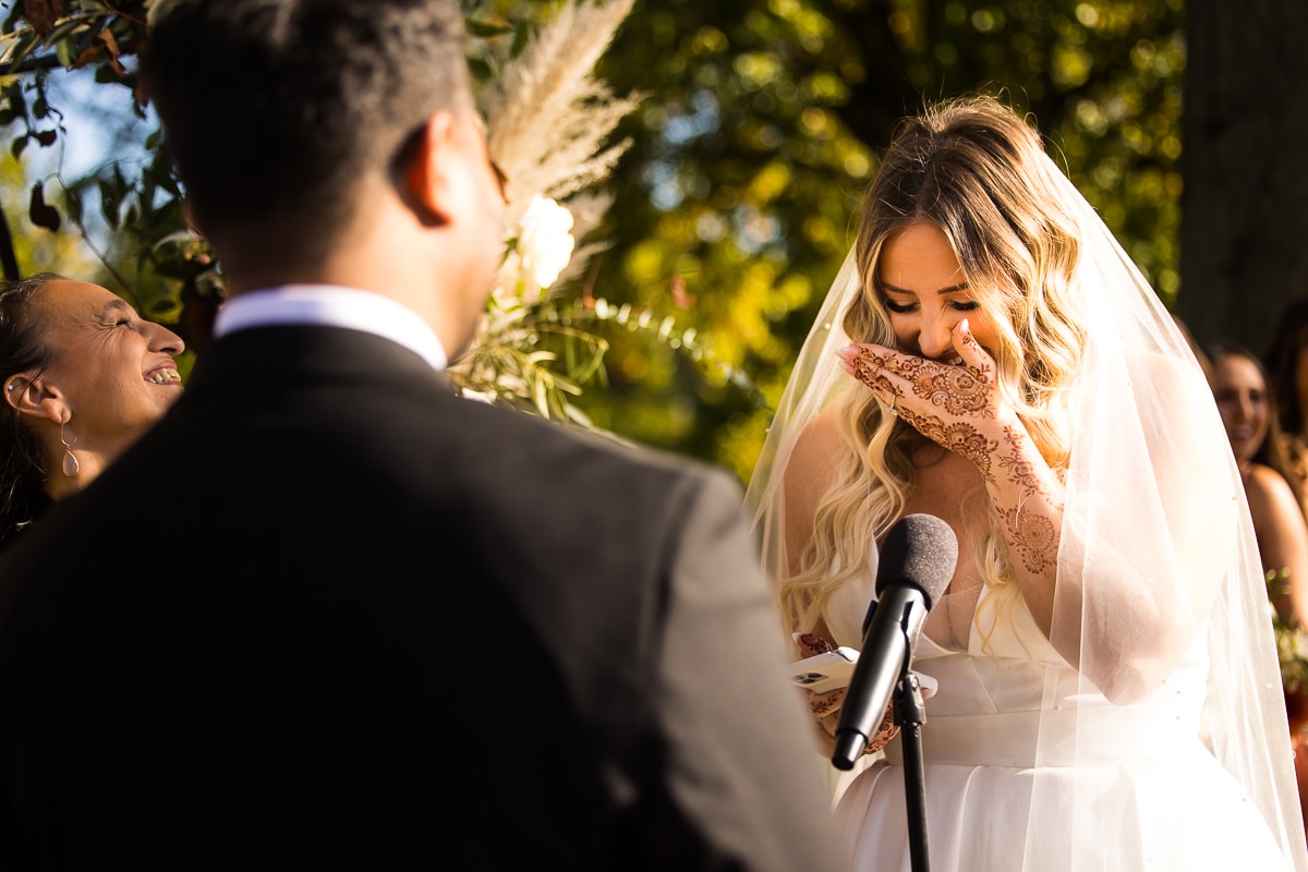 image of the bride laughing and covering her mouth with her henna decorated hand as she reads her vows to her husband during their Ashcombe Mansion wedding ceremony 