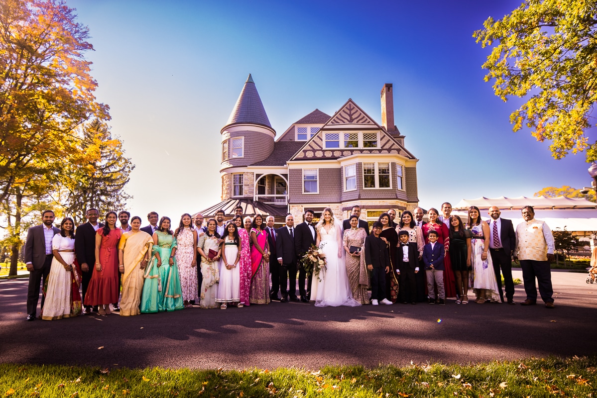 Wedding photographer, lisa rhinehart, captures the bride and groom with their whole family in a large portrait with the vibrant blue sky, the mansion, and gorgeous fall leaves behind them