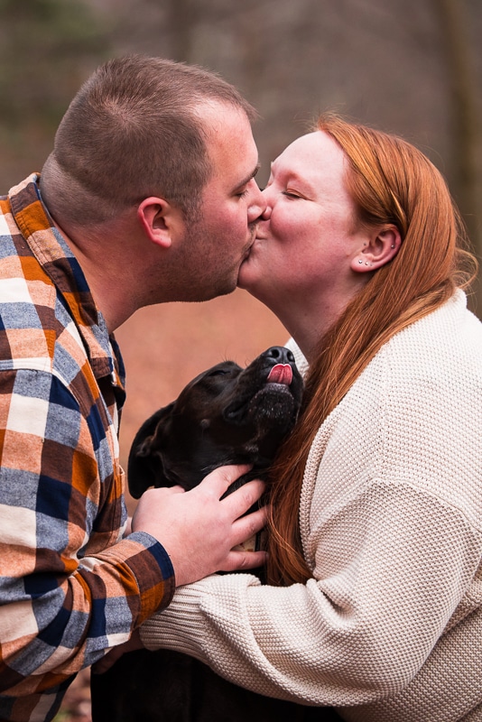 Image of the couple kissing as their dog looks up at them with it's tough out giving kisses too