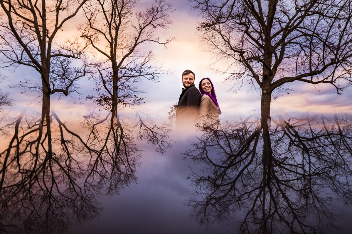 Unique, creative image taken by wedding photographer, Lisa Rhinehart, with the couple in the center of the image standing ack to back smiling at one another surrounded by trees with a blue, purple and orange sunset and the reflection of trees around them during this Murray Hill Engagement session in Virginia