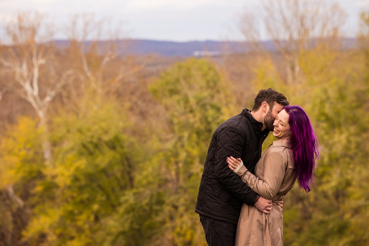 Image of the couple kissing on the cheek with a bunch of fall trees and leaves behind them at their wedding venue in Murray Hill, Virginia