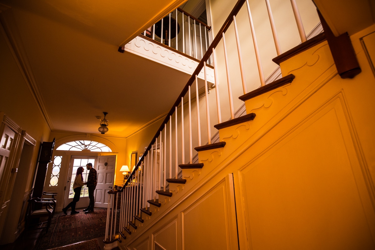 Dark, dramatic image of the couple standing in the door way with the large stair way in the image as well and the railing as a leading point to the couple inside Murray Hill, Virginia
