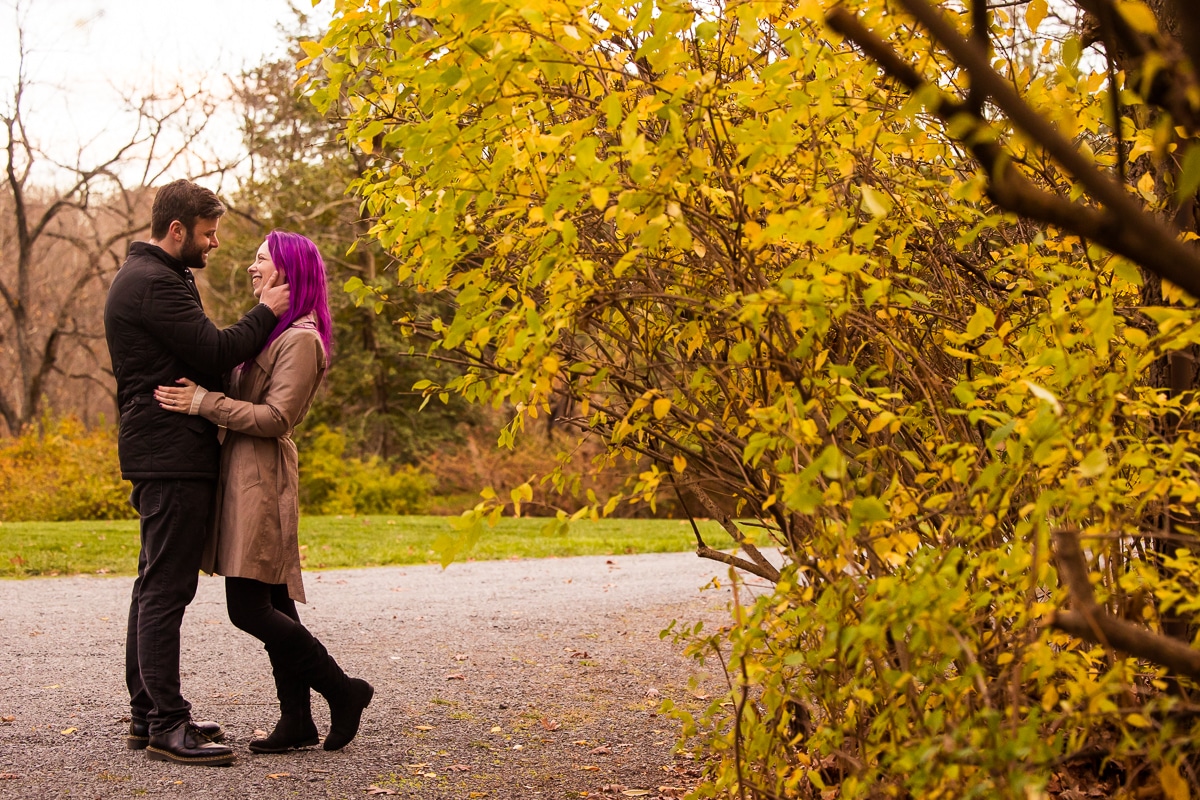 Traditional image of the couple smiling at one another and hugging as the fiance has his hand on her face surrounded by fall foliage during their Murray Hill Engagement session 