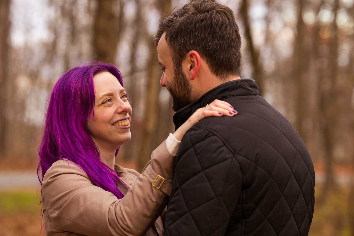 Image of the couple smiling at one another with the focus on the future bride smiling at her fiance as her purple hair pops against the fall foliage background in Virginia