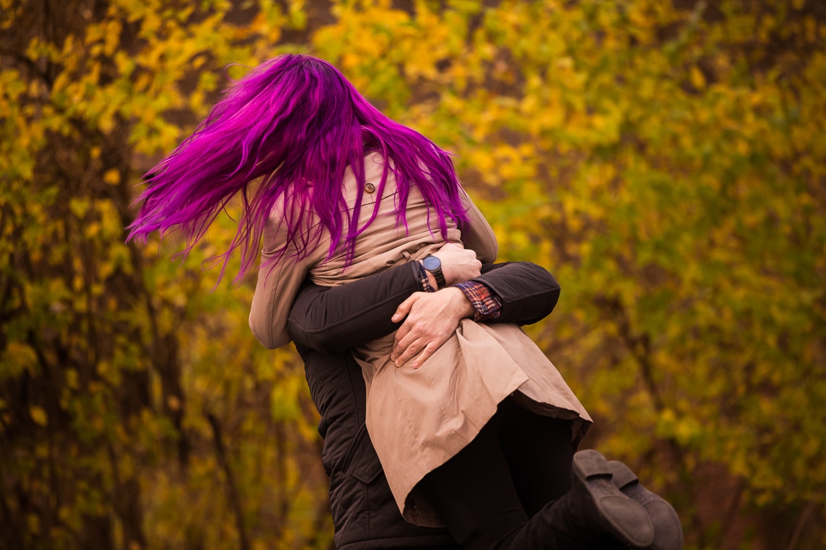 Image of the fiance hugging his future wife as he lifts her up and swings her around in the air as her vibrant, colorful purple hair flies in the wind during their engagement session at Murray Hill