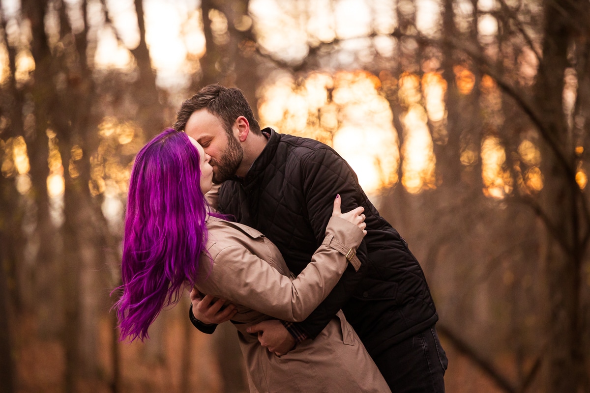 Image of the couple kissing as the sun sets in the trees behind them and the purple hair pops during this Murray Hill Engagement session