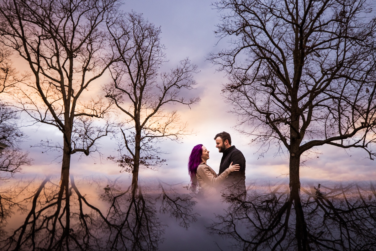 Unique, creative image taken by wedding photographer, Lisa Rhinehart, with the couple in the center of the image as they hug one another and smile while surrounded by trees with a blue, purple and orange sunset and the reflection of trees around them during this Murray Hill Engagement session in Virginia