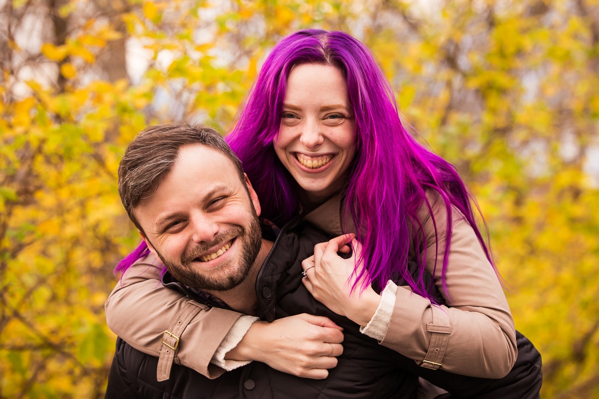 Image of the future bride with her vibrant, colorful purple hair on her fiance's back as they both smiling at the camera at Murray Hill in Virginia