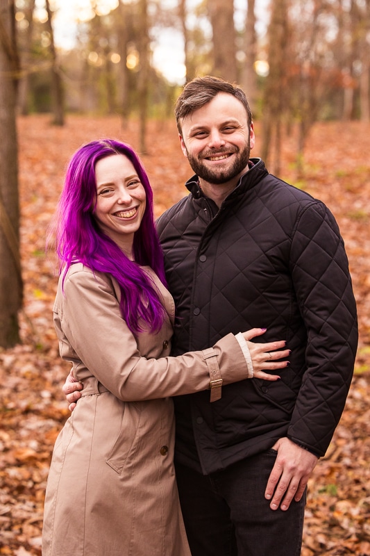Traditional portrait of the future husband and wife standing beside one another with her arm wrapped around him with leaves and trees filling the background behind them