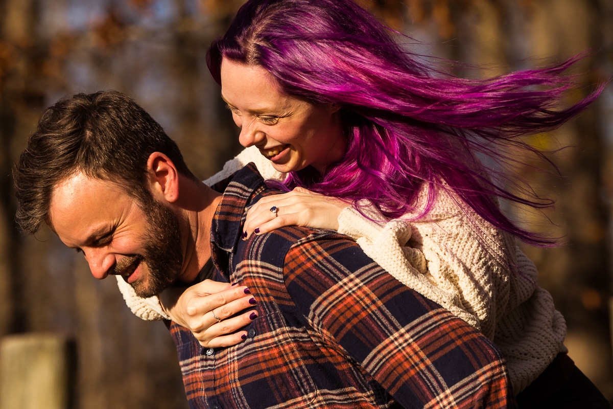 Murray Hill Engagement photographer, Lisa Rhinehart, captures the couple laughing and smiling as the future husband carries his future bride on his back with the vibrant, colorful purple hair blowing in the wind in Virginia