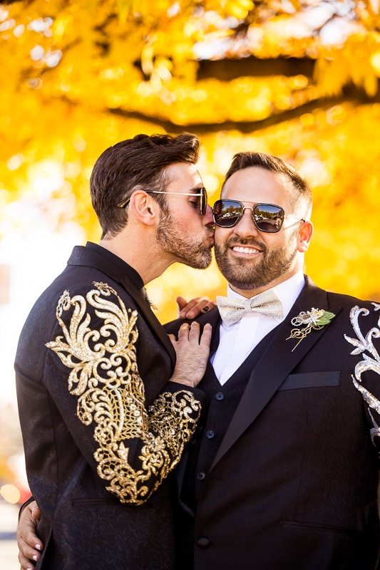 LGBT Wedding Photographer, Lisa Rhinehart, captures the grooms hugging and kissing each other on the cheek with a vibrant yellow, orange background of leaves behind them during their Phoenixville Foundry wedding in Philly